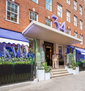 Property for sale by Auction An entrance to a stylish brick building with large windows, adorned with purple "34" signage above the main door. Blue flowers and cacti in planters embellish the entrance. A person in a beige suit and hat stands on the steps leading to the doorway. Presented by World of Auctions
