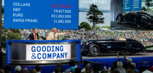 Property for sale by Auction An auctioneer and an assistant at a Gooding & Company event facilitate the sale of a vintage car. A large screen behind them displays currency conversions for the car's price. The audience eagerly watches and takes photos of the car on stage. Presented by World of Auctions