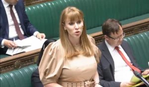 Property for sale by Auction A woman with auburn hair, dressed in a light beige dress, stands and speaks passionately at a parliamentary session. She is flanked by seated individuals in business attire, including a man in a black suit with a red tie holding a yellow document. Presented by World of Auctions