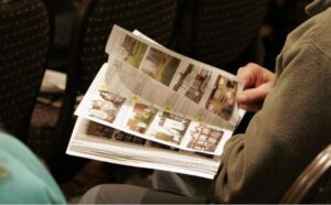 Property for sale by Auction A person sitting on a chair, leafing through a magazine or catalog featuring various property photographs and descriptions. The person is wearing a green jacket, and the surroundings suggest an indoor setting with more chairs visible in the background. Presented by World of Auctions