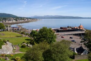 Property for sale by Auction A scenic coastal view of a small town with lush green parks, a pier with an orange-roofed building, and calm blue waters extending to distant mountains under a clear blue sky. Nearby roads and houses line the shoreline, creating a picturesque landscape. Presented by World of Auctions