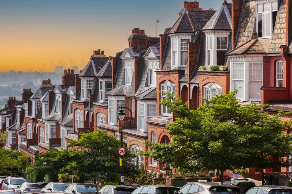 Property for sale by Auction A row of charming brick townhouses with gabled roofs and lush green trees lining the street. Cars are parked along the roadside under a clear sky transitioning from blue to orange at sunset. The scene reflects a peaceful suburban neighborhood. Presented by World of Auctions