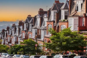 Property for sale by Auction A row of charming brick townhouses with gabled roofs and lush green trees lining the street. Cars are parked along the roadside under a clear sky transitioning from blue to orange at sunset. The scene reflects a peaceful suburban neighborhood. Presented by World of Auctions