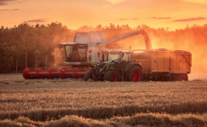 Property for sale by Auction A combine harvester and tractor work together in a wheat field at sunset, with the harvester transferring grain into the trailer pulled by the tractor. The golden light of the setting sun casts a warm glow over the scene, highlighting the dust and crops. Presented by World of Auctions