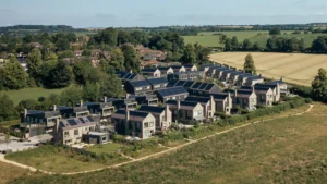 Property for sale by Auction Aerial view of a modern residential neighborhood with rows of houses, many equipped with solar panels on their rooftops. The area is surrounded by green fields and trees, with a small forested area in the background under a clear blue sky. Presented by World of Auctions