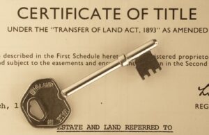 Property for sale by Auction A close-up of a Certificate of Title document with a metal key placed on top. The key has the word "ENGLAND" engraved on it and is positioned diagonally across the certificate, partially obscuring the text. The document references the Transfer of Land Act, 1893, illustrating why property auction lots sometimes fail. Presented by World of Auctions