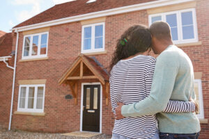 Property for sale by Auction A couple stands together with their arms around each other, looking at a two-story brick house with white-framed windows and a wooden porch. They appear to be facing the front door, suggesting they might be homeowners or prospective buyers. Presented by World of Auctions
