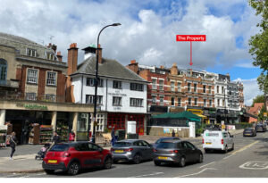 Property for sale by Auction Street view showing a row of buildings, including a four-story structure labeled "The Property." The scene includes cars parked and driving along the street, with shops and a tree on the right. The sky is partly cloudy. Presented by World of Auctions