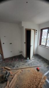 Property for sale by Auction A room under renovation with scattered construction materials. The floor is covered with sawdust and wooden planks. An open door leads to a small closet. There's a window with a grid design, and tools are lying on the ground. Walls are partially painted. Presented by World of Auctions