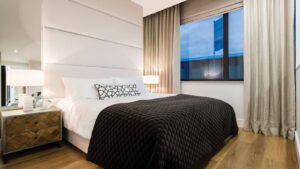 Property for sale by Auction A modern bedroom with a large bed featuring a white headboard, dark quilted bed cover, and geometric print pillow. A wooden nightstand with a lamp is beside the bed, and a large window with beige curtains lets in natural light. Presented by World of Auctions