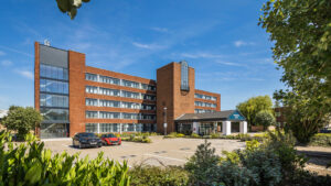 Property for sale by Auction A modern brick hotel with five stories, featuring a central glass atrium. The building is surrounded by greenery and a parking lot with a few cars. The sky is clear and blue, enhancing the bright atmosphere. Presented by World of Auctions