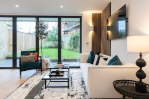 Property for sale by Auction Discover a modern living room in this Wimbledon new home for sale, with large glass doors opening to a garden. It features a white sofa with blue and black cushions, a dark armchair, a glass coffee table, ambient lighting, and a mounted TV on the wall. A monochrome rug completes the decor. Presented by World of Auctions