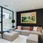 Property for sale by Auction A modern living room in a Wimbledon new home for sale features a large beige sectional sofa adorned with throw pillows. A vibrant landscape painting graces the dark accent wall. The room boasts a large window with an outdoor view and a small round side table, embodying elegant comfort. Presented by World of Auctions