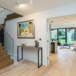 Property for sale by Auction A modern interior of a Wimbledon new home for sale features a wooden staircase with glass railings on the left. A console table with candles and a bowl sits beneath a colorful painting. In the background, a bright living room opens to a garden through large glass doors. Presented by World of Auctions