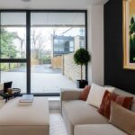Property for sale by Auction A modern living room in a Wimbledon new home for sale features a large window view. The room includes a light beige sofa adorned with white, brown, and rust pillows, a small round table with a book, and a framed landscape painting. Outside, the driveway and modern buildings complete the scene. Presented by World of Auctions