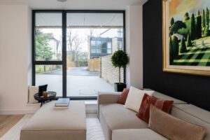 Property for sale by Auction A modern living room in a Wimbledon new home for sale features a large window view. The room includes a light beige sofa adorned with white, brown, and rust pillows, a small round table with a book, and a framed landscape painting. Outside, the driveway and modern buildings complete the scene. Presented by World of Auctions