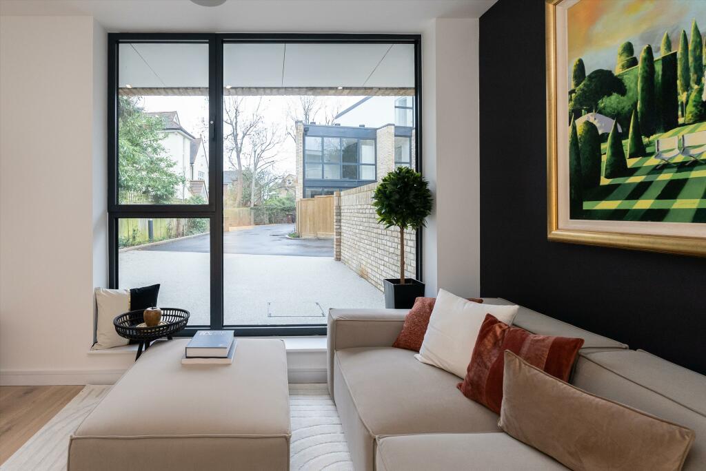 Property for sale by Auction A modern living room in a Wimbledon new home for sale features a large window view. The room includes a light beige sofa adorned with white, brown, and rust pillows, a small round table with a book, and a framed landscape painting. Outside, the driveway and modern buildings complete the scene. Presented by World of Auctions