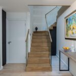 Property for sale by Auction Discover this Wimbledon new home for sale, boasting a modern hallway with a wooden staircase and sleek glass paneling. A side table showcases a candle, fruit bowl, and sculpture, while a vibrant painting on the white wall contrasts beautifully with dark wood accents. Presented by World of Auctions
