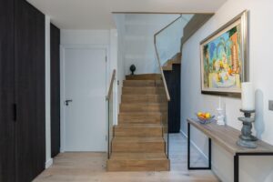 Property for sale by Auction Discover this Wimbledon new home for sale, boasting a modern hallway with a wooden staircase and sleek glass paneling. A side table showcases a candle, fruit bowl, and sculpture, while a vibrant painting on the white wall contrasts beautifully with dark wood accents. Presented by World of Auctions