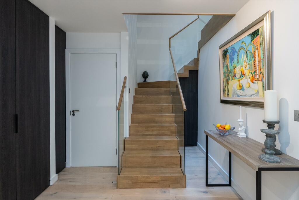 Property for sale by Auction Discover this Wimbledon new home for sale, boasting a modern hallway with a wooden staircase and sleek glass paneling. A side table showcases a candle, fruit bowl, and sculpture, while a vibrant painting on the white wall contrasts beautifully with dark wood accents. Presented by World of Auctions