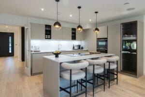 Property for sale by Auction A modern kitchen in this Wimbledon new home for sale features a large island with four cushioned bar stools. It boasts sleek, light-colored cabinets, a marble backsplash, and built-in ovens. Three pendant lights illuminate the island, and a fruit bowl adds charm to the countertop. Presented by World of Auctions