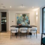 Property for sale by Auction Modern dining area in a Wimbledon new home for sale, featuring a sleek black table with six white, cushioned chairs. A large tropical painting adorns the wall. The space boasts wood flooring, a teal armchair, and expansive windows overlooking a rustic brick wall. Presented by World of Auctions