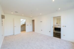 Property for sale by Auction Spacious empty room with white walls and beige carpet in this Wimbledon new home for sale. An open door reveals a staircase, while recessed lighting fixtures adorn the ceiling. A sliding door leads to a bathroom featuring a modern sink and dark tile flooring, all in a minimalist design. Presented by World of Auctions