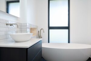 Property for sale by Auction This Wimbledon new home for sale boasts a modern bathroom with a minimalist design, featuring a white vessel sink on a dark vanity, an inviting large bathtub, and a frosted window that complements the light gray subway-tiled walls. Presented by World of Auctions