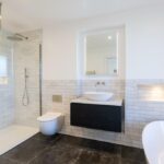 Property for sale by Auction A modern bathroom in a Wimbledon new home for sale boasts white subway tiles, a glass walk-in shower, wall-mounted toilet, black floating vanity with vessel sink, illuminated mirror, freestanding bathtub, and large floor tiles. Presented by World of Auctions