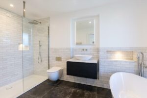 Property for sale by Auction A modern bathroom in a Wimbledon new home for sale boasts white subway tiles, a glass walk-in shower, wall-mounted toilet, black floating vanity with vessel sink, illuminated mirror, freestanding bathtub, and large floor tiles. Presented by World of Auctions
