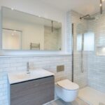Property for sale by Auction Discover a modern bathroom in this Wimbledon new home for sale. It boasts white subway tile walls, a glass-enclosed shower, a wall-mounted toilet, and a wooden vanity with sink. A large mirror sits above the vanity, enhanced by well-placed ceiling lights for optimal illumination. Presented by World of Auctions