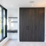 Property for sale by Auction A modern, empty room in a Wimbledon home for sale, featuring large glass doors on the left leading to a balcony. The space includes a dark wood wardrobe with two doors and a small built-in desk with shelves. White walls and light-colored carpet complete this inviting atmosphere. Presented by World of Auctions
