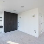 Property for sale by Auction A modern, empty room in a Wimbledon new home for sale features light gray carpet, white walls, and a dark wood built-in wardrobe. A small desk with shelves sits beneath two wall lights, while natural light streams in from a window. Doorways lead to other areas of this inviting space. Presented by World of Auctions