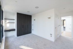 Property for sale by Auction A modern, empty room in a Wimbledon new home for sale features light gray carpet, white walls, and a dark wood built-in wardrobe. A small desk with shelves sits beneath two wall lights, while natural light streams in from a window. Doorways lead to other areas of this inviting space. Presented by World of Auctions