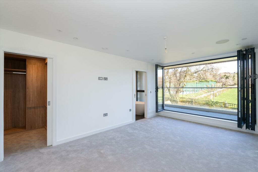 Property for sale by Auction This Wimbledon new home for sale boasts a spacious, modern bedroom with light-colored walls and carpet. Large bi-fold glass doors open to reveal a stunning view of the garden and trees. To the left, an open walk-in closet with wooden shelves adds both elegance and functionality. Presented by World of Auctions