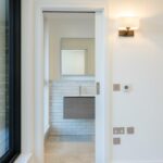 Property for sale by Auction Through the doorway of this Wimbledon new home for sale, you'll find a minimalist bathroom featuring a modern sink with a rectangular mirror above it. The white-tiled walls and wall-mounted light ensure the space is well-lit, while a large window partially visible to the left invites natural light in. Presented by World of Auctions