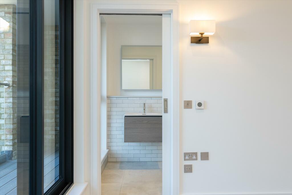 Property for sale by Auction Through the doorway of this Wimbledon new home for sale, you'll find a minimalist bathroom featuring a modern sink with a rectangular mirror above it. The white-tiled walls and wall-mounted light ensure the space is well-lit, while a large window partially visible to the left invites natural light in. Presented by World of Auctions