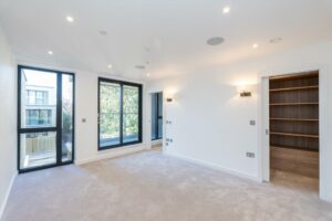 Property for sale by Auction A bright, empty room in a Wimbledon new home for sale features light beige carpet, white walls, and large windows that flood the space with natural light. Two built-in wall lamps add to the gentle ambiance. A door opens to a wooden closet with shelves on the right. Presented by World of Auctions