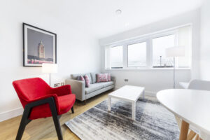 Property for sale by Auction A bright living room with a gray sofa, red armchair, and white coffee table on a gray rug. A large window provides natural light, and there's a floor lamp in the corner. A framed picture of a clock tower hangs on the wall. Presented by World of Auctions