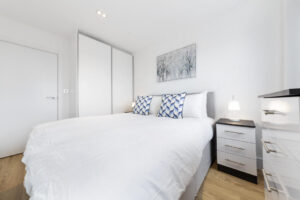 Property for sale by Auction A modern bedroom with a neatly made double bed. The bed has white bedding and two patterned blue and white pillows. There's a bedside table with a small lamp and a wardrobe with sliding doors. A framed artwork hangs on the white wall above the bed. Presented by World of Auctions