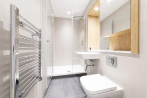 Property for sale by Auction Modern, minimalist bathroom with a glass shower, wall-mounted sink, white toilet, and tall chrome towel radiator. Light wooden accents and neutral tones create a clean and spacious feel. Presented by World of Auctions