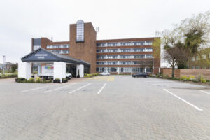 Property for sale by Auction A modern brick building with several floors labeled "Arrowhead House" at the entrance. The structure features large windows and a glass entrance. The foreground shows a spacious parking lot with a few parked cars and neatly landscaped greenery. Presented by World of Auctions