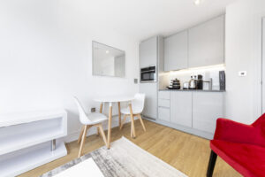 Property for sale by Auction A modern, compact kitchen and dining area with light grey cabinets, a small round table, two white chairs, and a red armchair. The room features wood flooring and a white wall with a mirror, creating a bright and airy atmosphere. Presented by World of Auctions