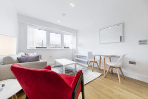 Property for sale by Auction A modern, bright living and dining area with white walls. It features a red armchair, a gray sofa, a wooden dining table with white chairs, and a large window letting in natural light. A rug and a wall mirror complete the decor. Presented by World of Auctions