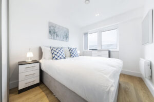 Property for sale by Auction A bright, modern bedroom with a large bed dressed in white linens and blue-patterned pillows. There are two bedside tables with lamps, a framed artwork above the bed, and a window letting in natural light. The floor is wooden. Presented by World of Auctions