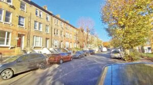 Property for sale by Auction A sunny street with several brick townhouses lined along one side. Cars are parked on both sides of the street. Trees with autumn leaves are on the right, under a clear blue sky. Presented by World of Auctions