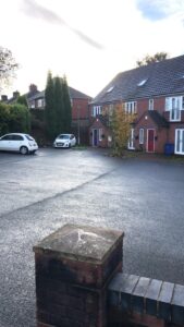 Property for sale by Auction A residential street with a parking area featuring two cars, a white one and a partially visible dark-colored one. Brick houses with gabled roofs and colorful doors line the background, and a small tree is in the foreground. Presented by World of Auctions