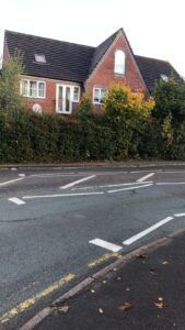 Property for sale by Auction A two-story brick house with a dark gray roof is partially obscured by a hedge. It's situated behind a road with painted traffic lines and a yellow curb. The hedge has green leaves with touches of autumn colors. Presented by World of Auctions