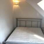 Property for sale by Auction A room with a metal-framed bed, a white mattress, and a sconce light on the slanted wall above the bed. There is natural light coming through a skylight on the right side. Walls and ceiling are painted white. Presented by World of Auctions
