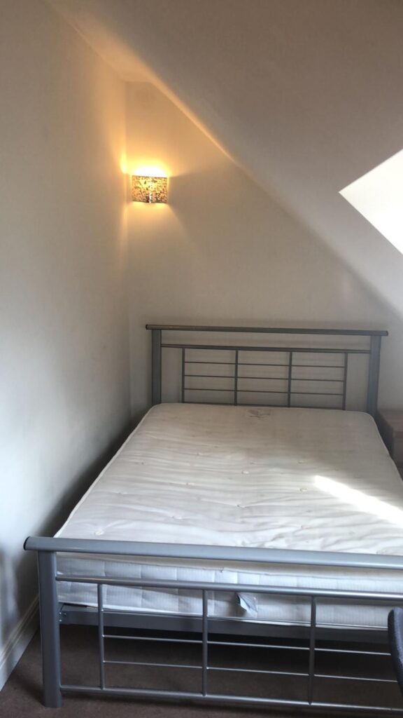 Property for sale by Auction A room with a metal-framed bed, a white mattress, and a sconce light on the slanted wall above the bed. There is natural light coming through a skylight on the right side. Walls and ceiling are painted white. Presented by World of Auctions